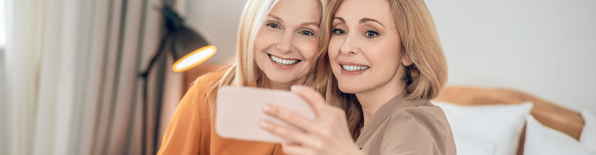 Two smiling blonde women sitting on the bed and making selfie on a smartphone