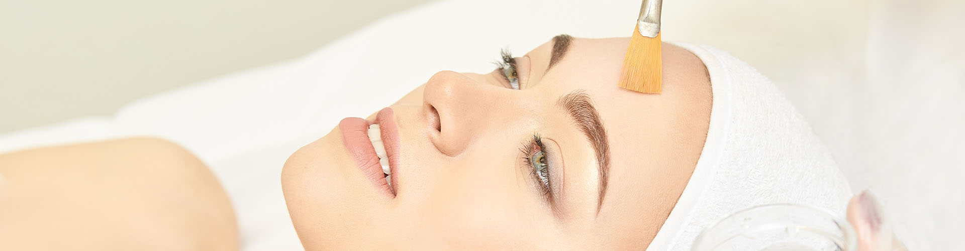 Person lying down with a white headband having a facial treatment applied to their forehead with a brush.