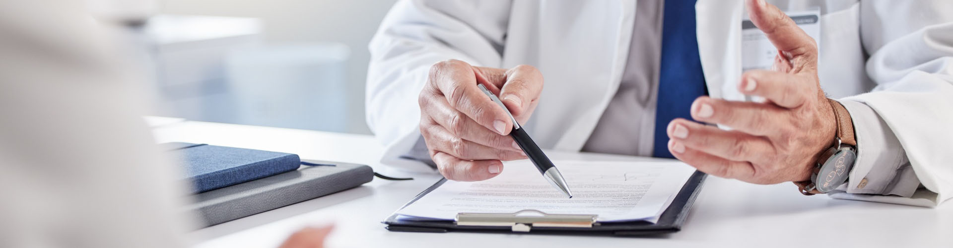 A doctor in a white coat sits at a desk, holding a pen and explaining information on a clipboard to a patient.